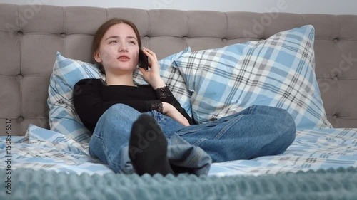 Young Woman Teen Girl Relaxing In Comfortable Casual Clothes Lying On Bed With Soft Pillows, Talking On Smartphone With Peaceful Expression While Leisure At Home