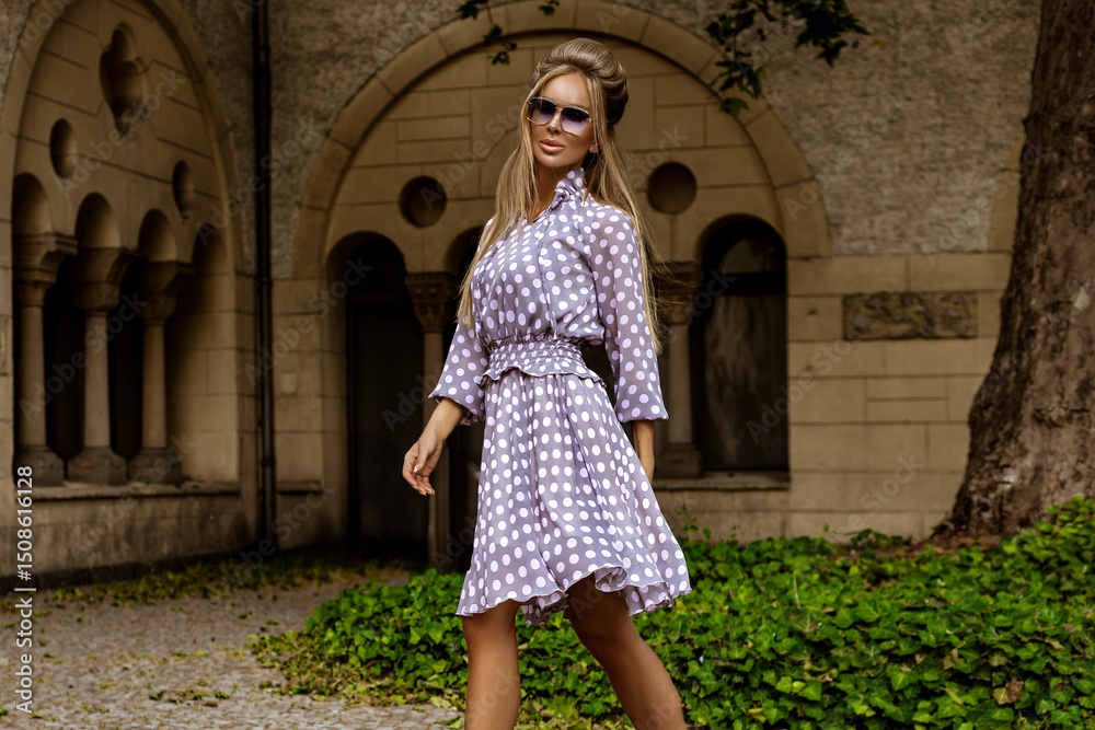 Elegant woman in long polka dot summer dress and fashionable high heels poses sensually in the city on a sunny summer day. Summer Fashion. Elegant woman in long polka dot summer dress and fashionable high heels poses sensually in the city on a sunny summer day. Summer Fashion.