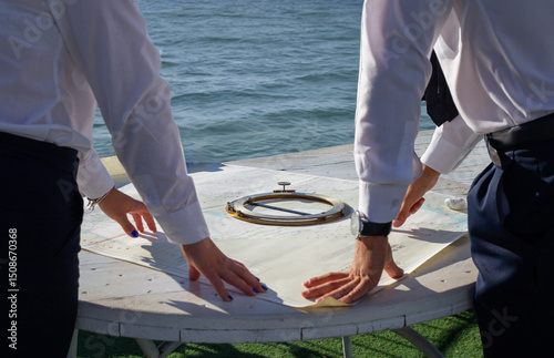 Two navigators in white shirts examine a nautical map on a white table, with a compass in the foreground and the sea in the background