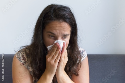 Sick caucasian woman sitting on sofa, wiping runny nose with tissue during illness, showing viral infection symptoms