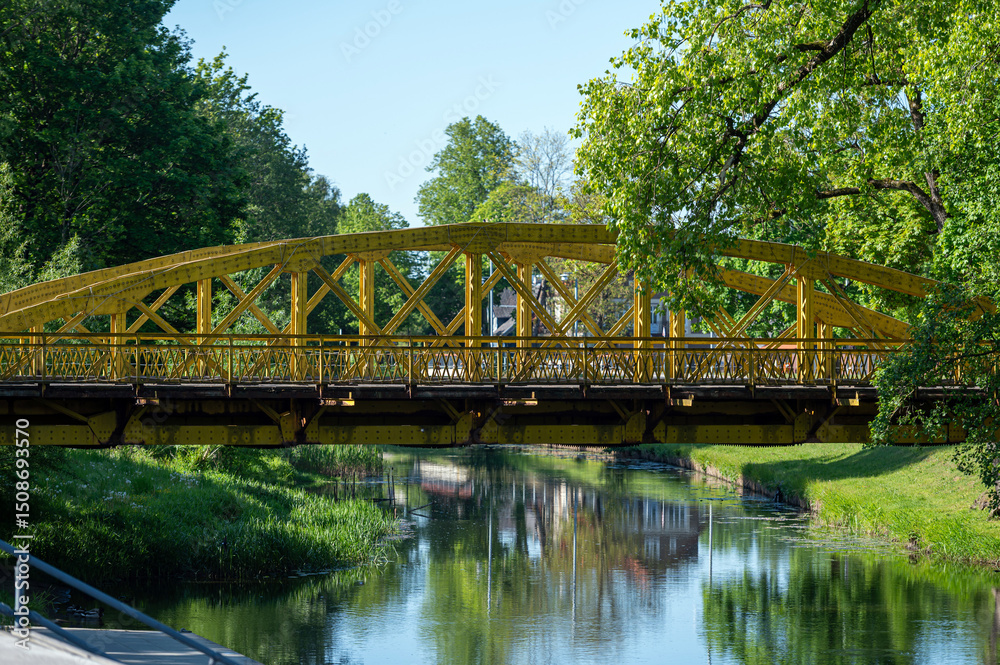 Obraz premium Colorful bridge spanning a calm river surrounded by lush greenery on a sunny day
