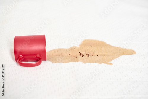 Red mug lies on side, spilling coffee onto white fabric. Light coffee stain spreads, creating splash effect. Contrasting colors highlight spills prominence