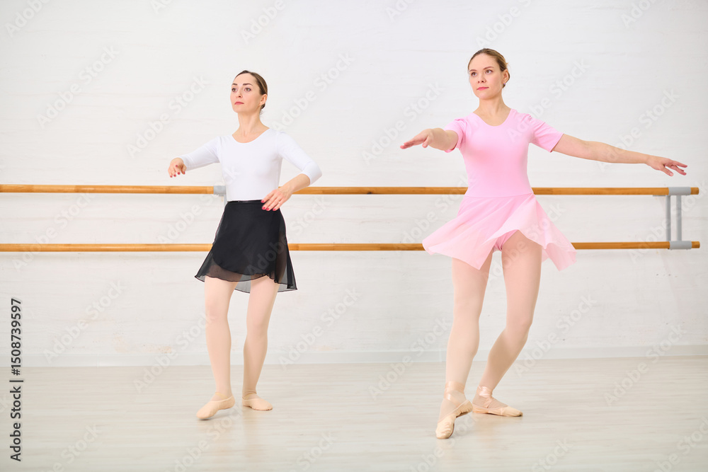 Naklejka premium Young female ballerinas with light hair practicing at barre in bright studio. One wears black skirt while other wears pink dress. Natural light enhances calm atmosphere