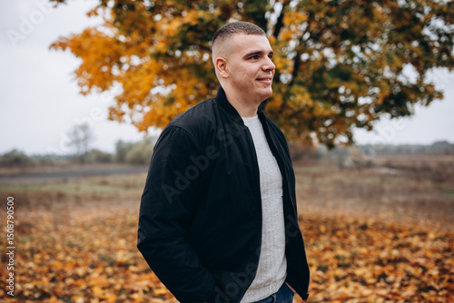 Smiling young man standing outdoors in an autumn park. He is wearing a black jacket and jeans, posing confidently in front of a tree with vibrant orange and yellow leaves. Casual and cheerful mood.