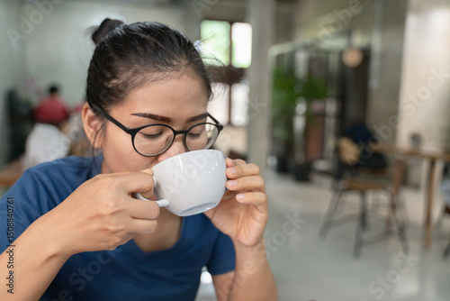 Young woman with glasses enjoying a warm beverage from a white cup in a cozy modern cafe, surrounded by a calm indoor atmosphere.
