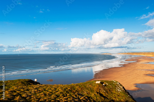 A scenic view from a grassy cliff overlooking a vast beach and ocean. The tide is out, revealing a wide expanse of sand, and the sky is a mix of blue and fluffy white clouds.