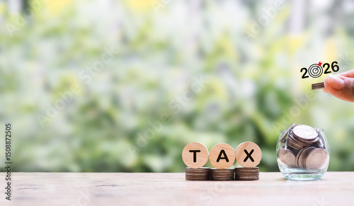 Hand putting coin into glass bottle and wooden block with word TAX on stack of coins. The concept of saving money for payment tax in year 2026.