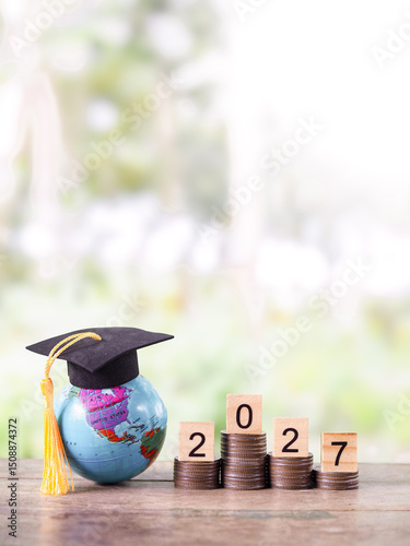 Earth globe with graduation hat, Wooden block with number 2027 on stack of coins. The concept of saving money for education, student loan, scholarship, tuition fees in year 2027.