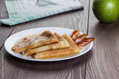 Photo of crepes, thin pancakes, British pancakes with apple jam on the white plate on wooden table with kitchen napkin and apple in background.