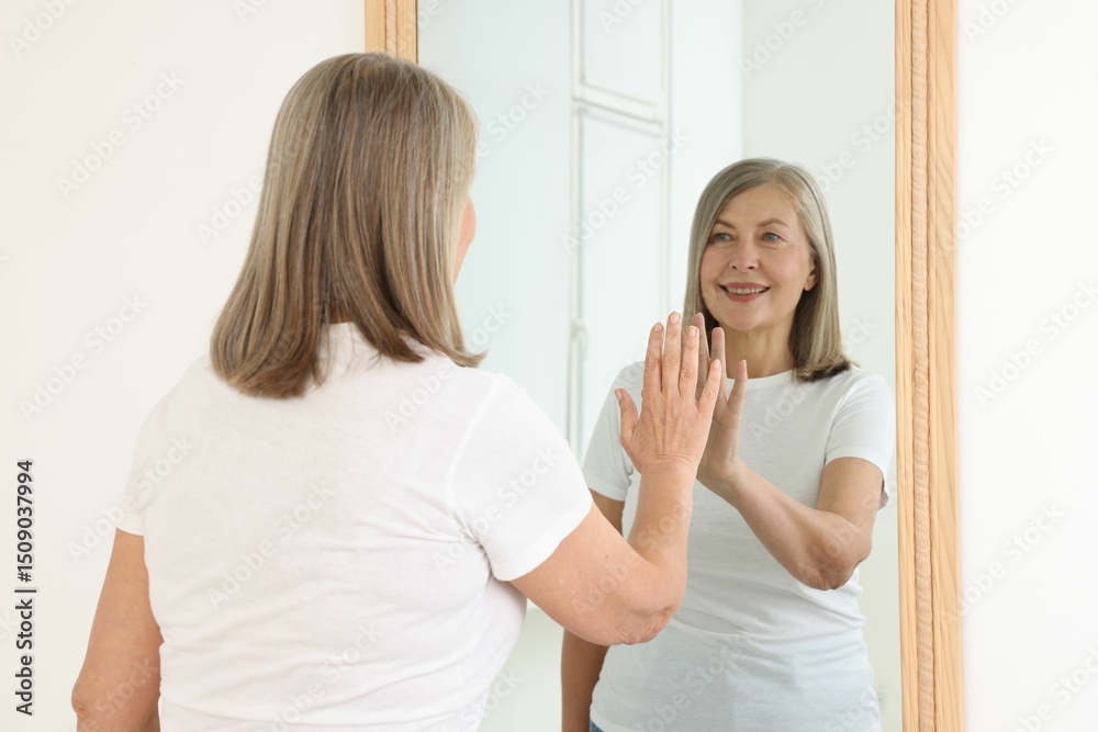Fototapeta premium Beautiful senior woman looking at herself in mirror indoors
