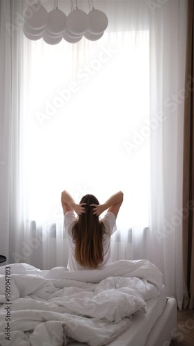 A young woman sits on the edge of the bed, stretching after waking up on a sunny morning
