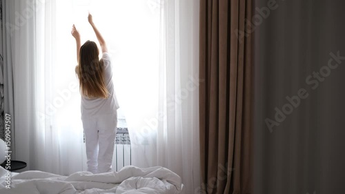 A young woman in cozy homewear shuts the bedroom window to keep out the chilly morning air, then tidies up the bed in soft daylight