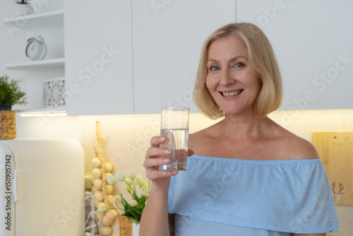 A smiling woman holds a refreshing glass of water surrounded by fresh vegetables in a stylish kitchen