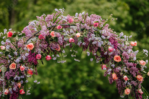 Close-up of a lush wedding floral arch made of lilacs, roses, and purple blossoms, with green foliage and soft natural background.. photo