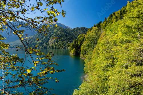 On a warm autumn day, a picturesque view of the famous Lake Ritsa opens up, visible through the foliage of the trees of the Abkhazian mountain range.