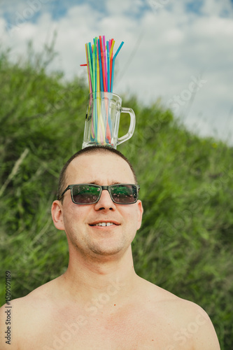 A guy in sunglasses holds a beer glass with straws on his head. Summer, vacation, holiday concept