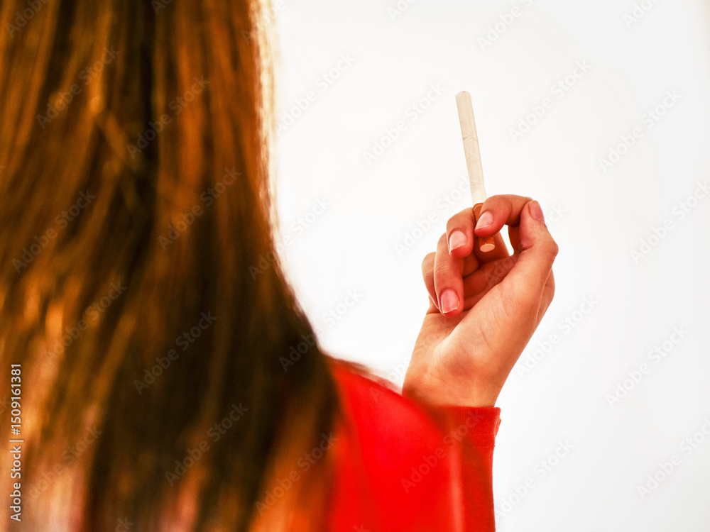 Obraz premium A young girl holding a cigarette in her hand with stylish nails. Concept of addiction and the dangers of smoking. The model has red color shirt and there is light color background. Bad habit theme.