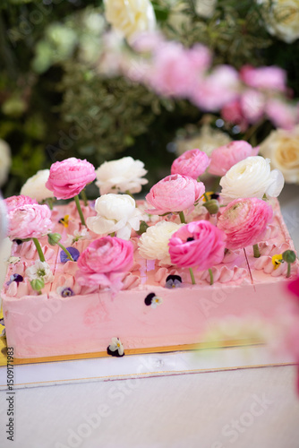Rectangular pink frosted cake decorated with fresh pink and white ranunculus flowers and edible blossoms on white table. Studio food photography. Wedding dessert and floral styling concept. Design for