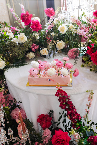 Pink rectangular cake decorated with white and pink ranunculus flowers placed on round white table surrounded by floral arrangements of roses and greenery. Indoor wedding celebration setup. Wedding de