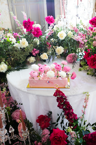 Pink rectangular cake decorated with white and pink ranunculus flowers placed on round white table surrounded by floral arrangements of roses and greenery. Indoor wedding celebration setup. Wedding de