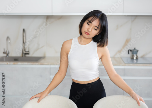 Upset young Asian woman standing in melancholic pose against background of white kitchen cabinet