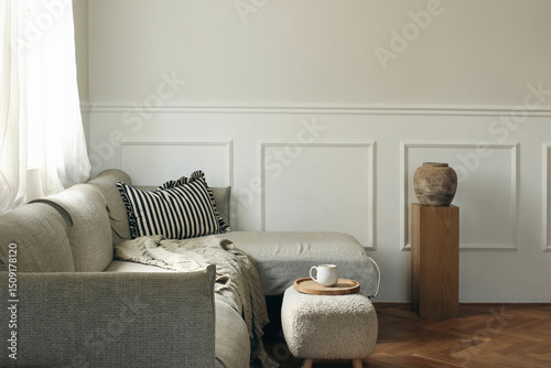 Elegant living room decor. Modern Scandinavian interior design. Cup of tea, coffee on tray, foot stool. Grey sofa, wooden pedestal with textured vase. Beautiful classic wall molding. Empty copy space.