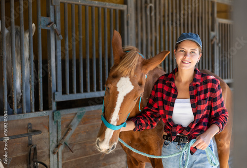 Young woman leading tethered brown horse out of stable