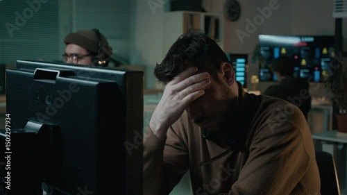 Medium shot of dismayed Caucasian scam caller sitting in illegal call center, staring at computer screen after failing to swindle victim with fake investment, feeling tired and rubbing face