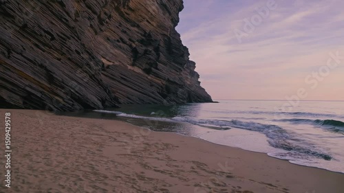 Tranquil ocean waves washing sandy beach near rocky cliff, casting soft twilight glow across peaceful coastal landscape