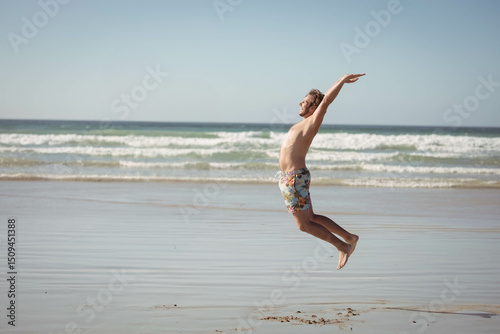 Man jumping above wet sand near shoreline at low tide, wearing colorful floral swim trunks
