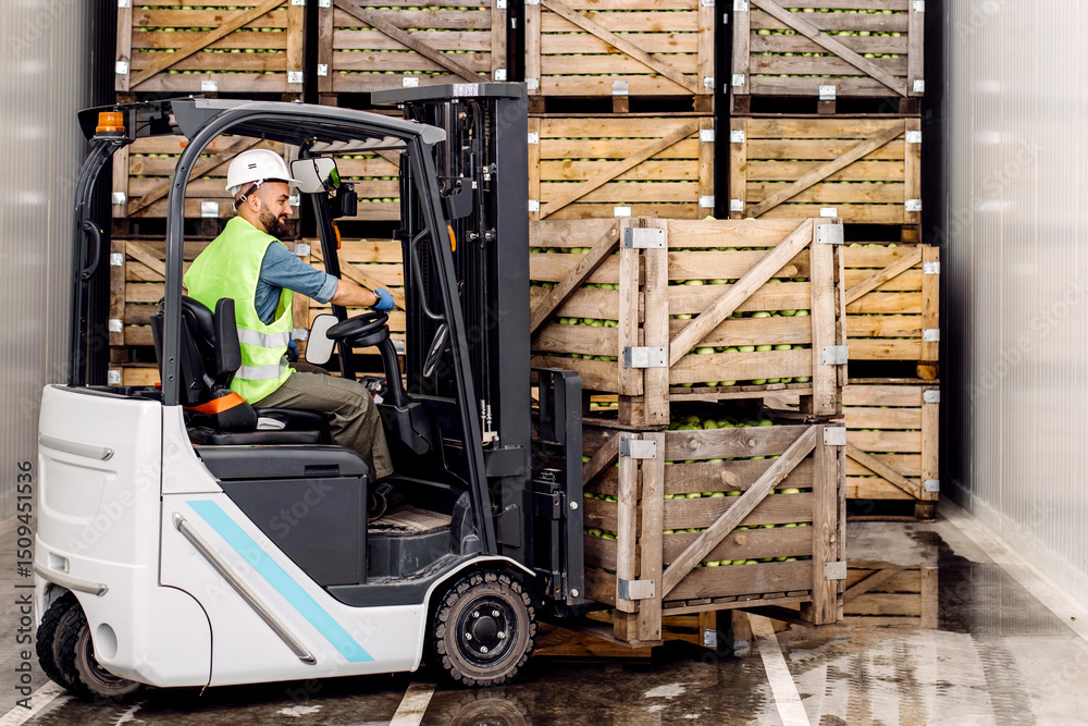 © Prostock-studio - Modern warehouse, work on forklift, industry and organic agribusiness. Smiling millennial man in helmet in forklift lifts up green apples in container on shelves with many wooden crates, free space
