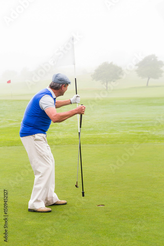 Senior golfer standing on manicured putting green, lifting flagstick from hole in misty morning
