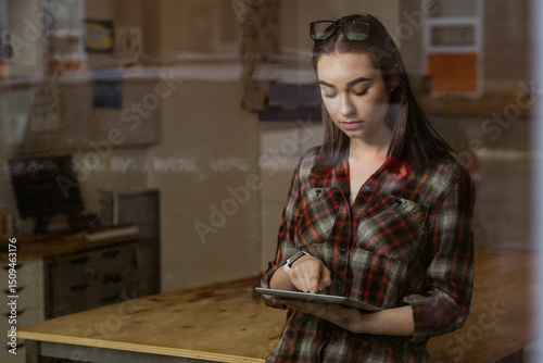 Woman in twenties in plaid shirt standing at office desk using tablet with pinboard, copy space photo