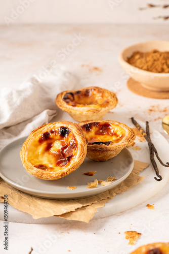 Three Pastel de Nata, Portuguese custard tarts, are presented on a plate with cinnamon and vanilla beans.