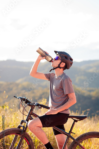 Cyclist resting and drinking water after a trail ride. He is sitting on his mountain bike and wearing a protective helmet. Concept of hydration, outdoor activity, and healthy lifestyle.
