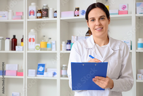 Smiling female pharmacist in white coat holding clipboard and pen in pharmacy...