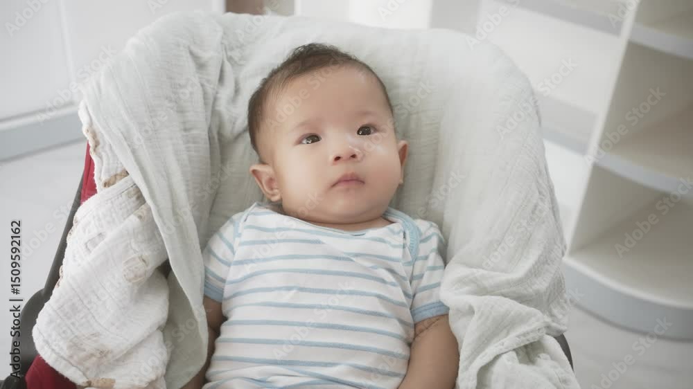 Asian curious 3-month-old baby infant with dark hair looks upwards, nestled comfortably in a red and black stroller indoors, wrapped in a light blanket. Asian curious 3-month-old baby infant with dark hair looks upwards, nestled comfortably in a red and black stroller indoors, wrapped in a light blanket.