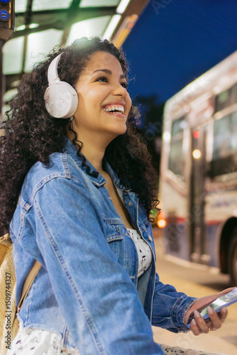 Independent young latin woman, smiling, listening music with headphones, waiting for the bus alone at night