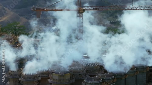 Cylindrical cooling towers release heavy white steam beneath large construction crane at Barrick Gold’s gold and silver mining complex near Cotuí, Dominican Republic. Slow pan left aerial photo