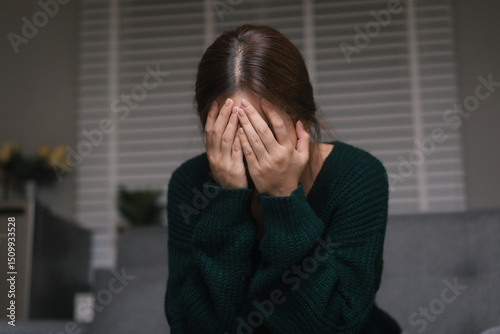 A young woman, who appears to be of Asian descent, sits curled on the floor with a melancholic expression, leaning against gray sofa, conveying feelings of loneliness and sadness in dimly lit room.