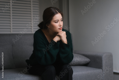 Canvas Print A young woman, who appears to be of Asian descent, sits curled on the floor with a melancholic expression, leaning against gray sofa, conveying feelings of loneliness and sadness in dimly lit room