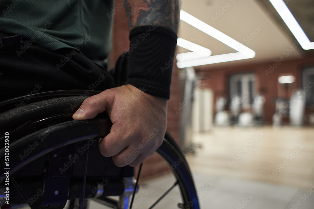 Obraz premium Close-up of tattooed arm resting on wheelchair handle with modern gym equipment blurred in background suggesting active and accessible fitness environment