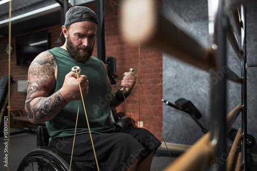 Man in wheelchair demonstrating strength while exercising with resistance cables at gym setting. Muscular build and tattoos visible, working out with determination