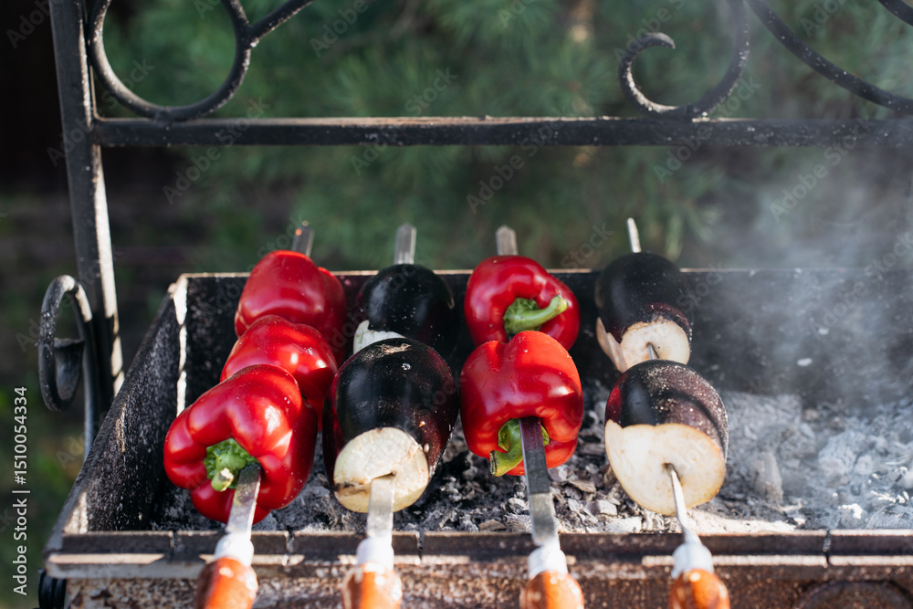 Obraz premium Close-up of red bell peppers and eggplants grilling on skewers over hot coals in an outdoor metal barbecue, with smoke rising in natural daylight.