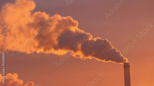 At sunset, a factory chimney expels dense smoke, contrasting the vivid sky and