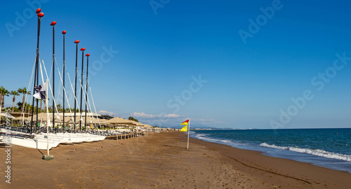 Fototapeta Naklejka Na Ścianę i Meble -  Panoramic view of a quiet beach with sailboats, empty loungers, sunshades, and signal flags on a sunny April day. Sorgun, Antalya, Mediterranean.