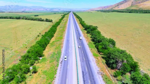 Drone shot of a highway cutting through green fields on a sunny day. Cars drive along the road stretching into the distance.