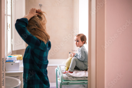 A mother and her young daughter spend time together in the bathroom during their morning routine. The mother fixes her hair while the child sits nearby. photo