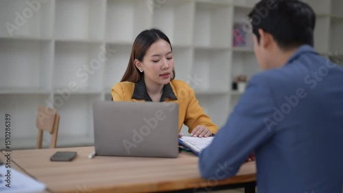 A woman and a man are sitting at a table with a laptop and a book. The woman is pointing at the laptop, possibly showing the man something on the screen. They are engaged in a conversation