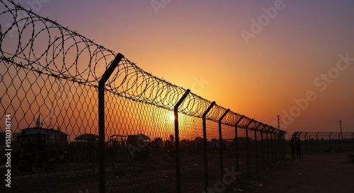 A barbed wire fence silhouetted against a vibrant sunset with orange and yellow hues in the background